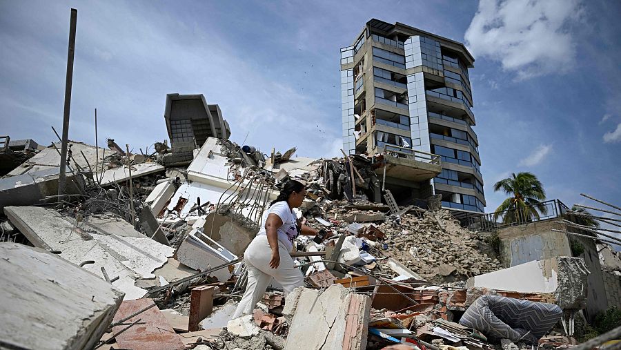 Mujer camina sobre escombros de edificios colapsados en un paisaje urbano devastado. Al fondo, un edificio moderno y una palmera se alzan entre la destrucción.