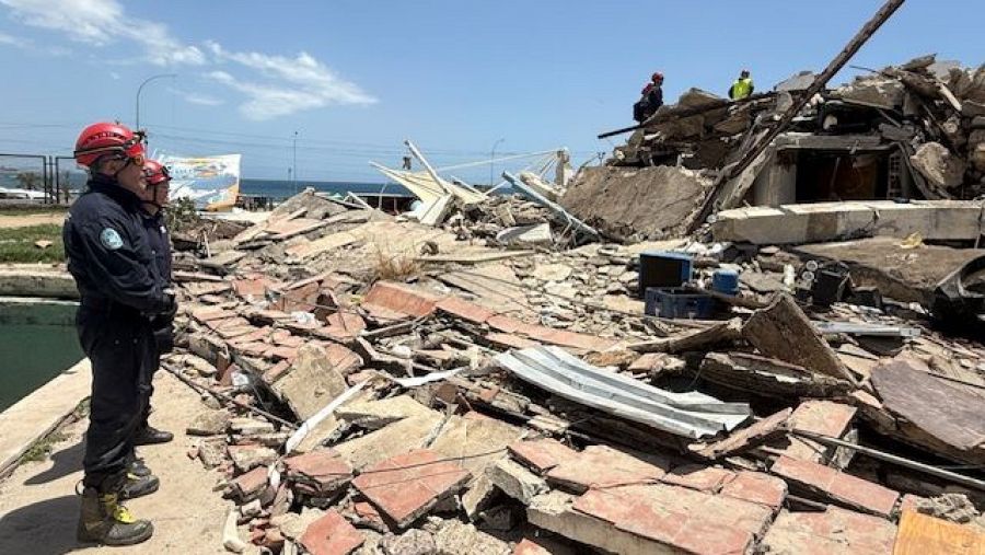 Bomberos españoles con cascos rojos y uniformes azules observan una zona de escombros y destrucción en La Guaira, Venezuela, tras un terremoto.