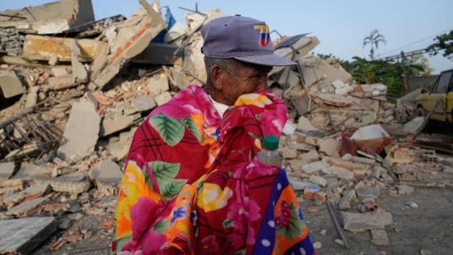 Hombre mayor con gorra de Venezuela y manta colorida en un paisaje de escombros, sosteniendo una botella de agua. Expresión de cansancio y angustia.