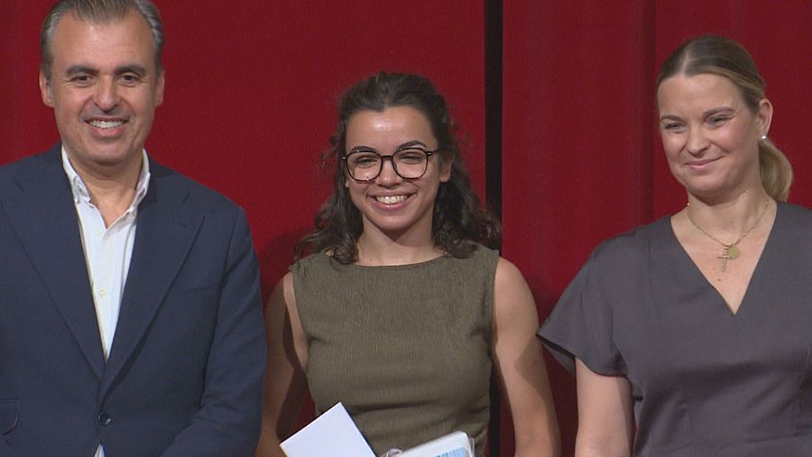 Miquel Ensenyat Riutort, Catalina Cladera Cifre y una joven sonríen en un evento formal. La joven sostiene un diploma y un folleto. Fondo rojo.