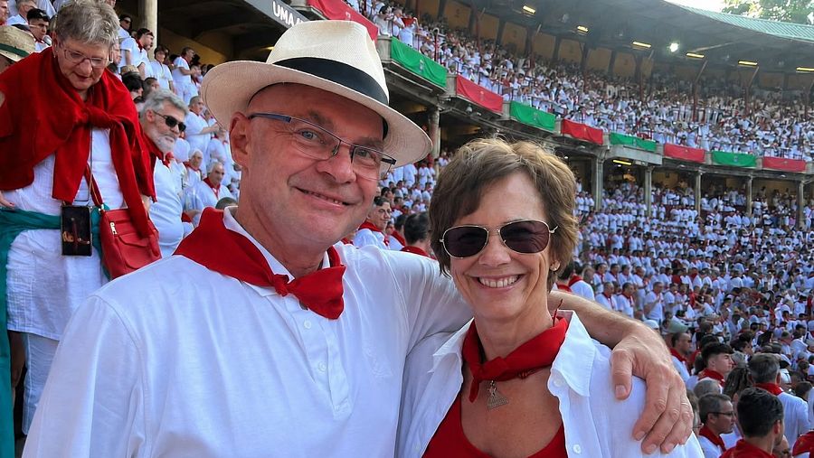 María y Hermud, en primer plano con atuendo de San Fermín, sonríen en una plaza de toros abarrotada con banderas de Navarra al fondo.