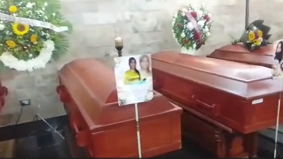 Cuatro ataúdes de madera oscura en una sala de velatorio, con una fotografía de dos mujeres, arreglos florales y una vela. Ambiente de luto y recuerdo.