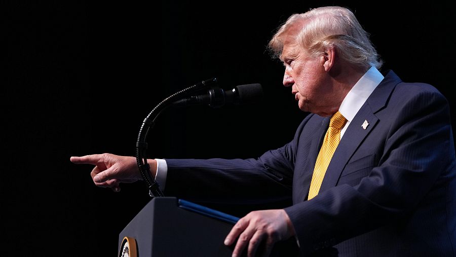 Donald Trump, de perfil, habla desde un podio con el brazo extendido y el dedo índice apuntando. Viste traje oscuro, camisa blanca y corbata amarilla. Fondo negro.