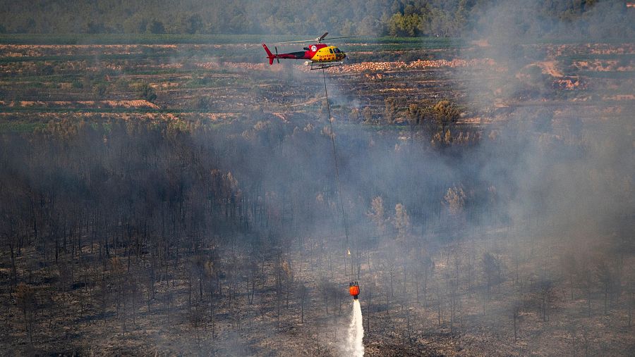 Helicóptero de extinción de incendios en acción, liberando agua sobre un paisaje quemado con humo denso. El bambi bucket naranja es visible.