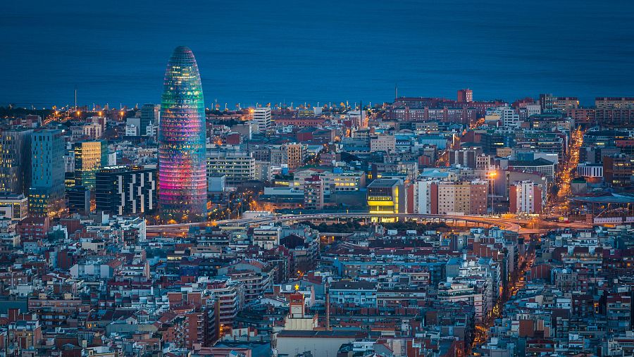 Vista aérea nocturna de Barcelona, con la Torre Glòries brillando en múltiples colores sobre un mar de luces urbanas.