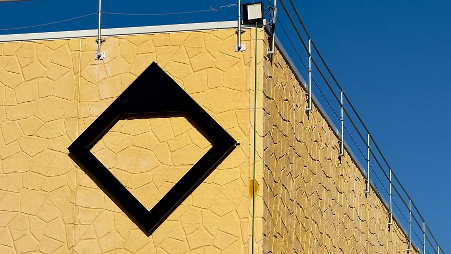 Logo negro en forma de diamante con parte superior rellena y contorno grueso, sobre pared amarillo-beige rugosa de edificio. Incluye barandilla, foco y cables bajo cielo azul.