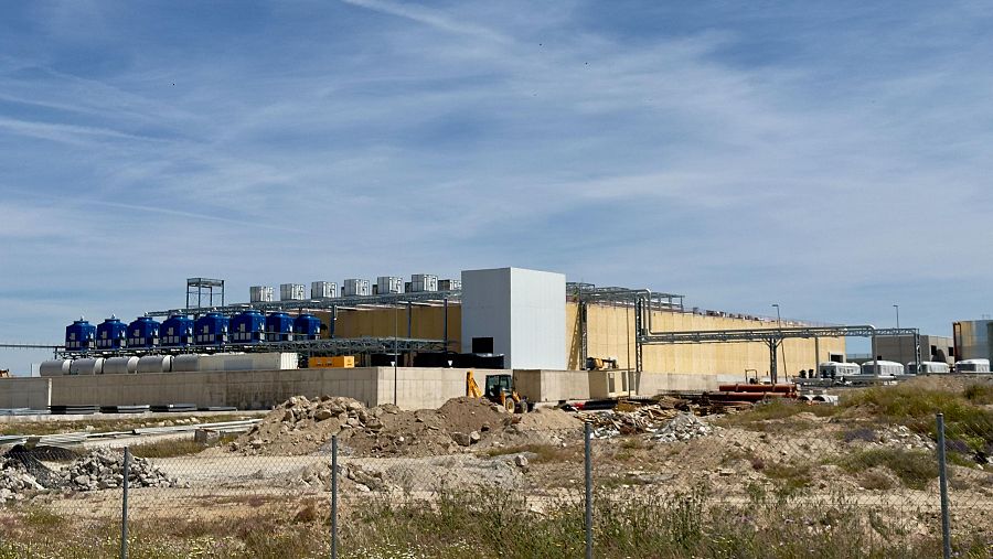 Instalación industrial moderna con tanques azules, edificio beige y blanco, terreno árido con retroexcavadora y valla metálica.