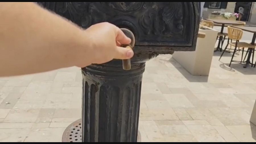 Mano girando pomo de bronce en fuente clásica de metal oscuro, con rejilla en el suelo y mobiliario de terraza al fondo.