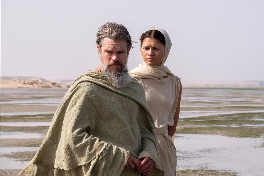 Matt Damon y Zendaya en un paisaje desértico, ambos con expresiones serias y vestimentas holgadas. El entorno árido y el cielo despejado crean una atmósfera cinematográfica.