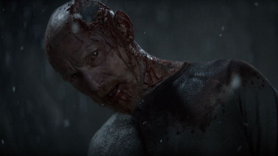 Primer plano de un hombre con heridas graves y sangre cubriendo su rostro y torso. Ojos abiertos y penetrantes en un ambiente oscuro y desolador.