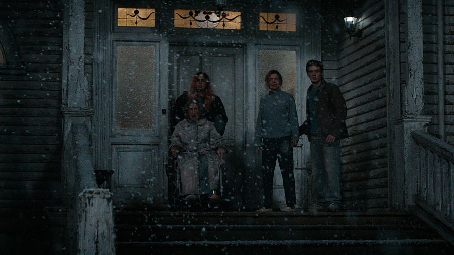 Cuatro personas, una de ellas en silla de ruedas, se congregan en la entrada nevada de una casa antigua y descuidada, transmitiendo una atmósfera de tensión y preocupación.