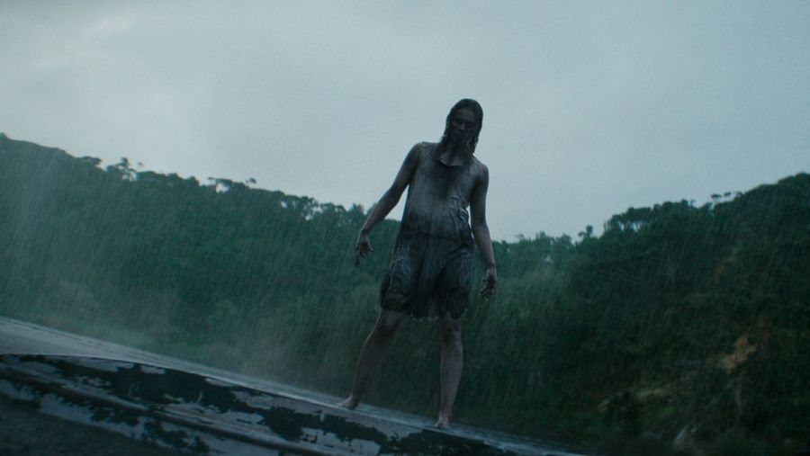 Mujer descalza y empapada bajo intensa lluvia, con vestido claro desgastado y expresión perturbada, en un entorno boscoso y sombrío.
