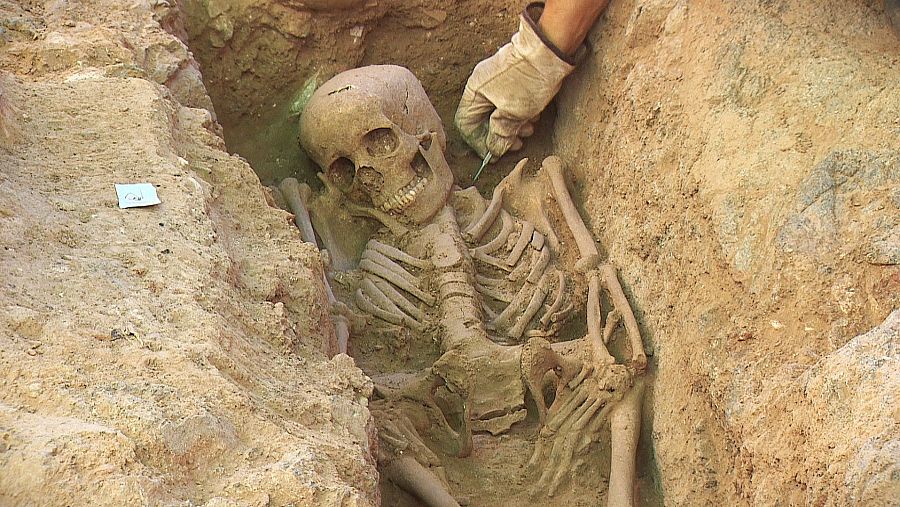Arqueólogo con guante beige usa herramienta fina sobre esqueleto visigodo en excavación en Mérida, Extremadura. Huesos terrosos en fosa de tierra, cráneo y extremidades visibles.