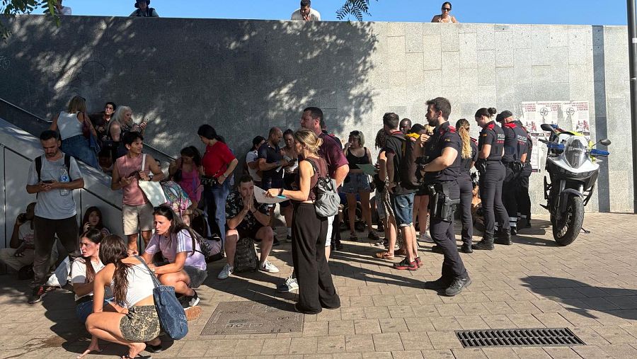 Multitud diversa y policía en escena urbana soleada. Agentes de Mossos d'Esquadra con motocicleta supervisan a personas en escaleras y suelo. Un hombre con barba y instrumento de cuerda presente.