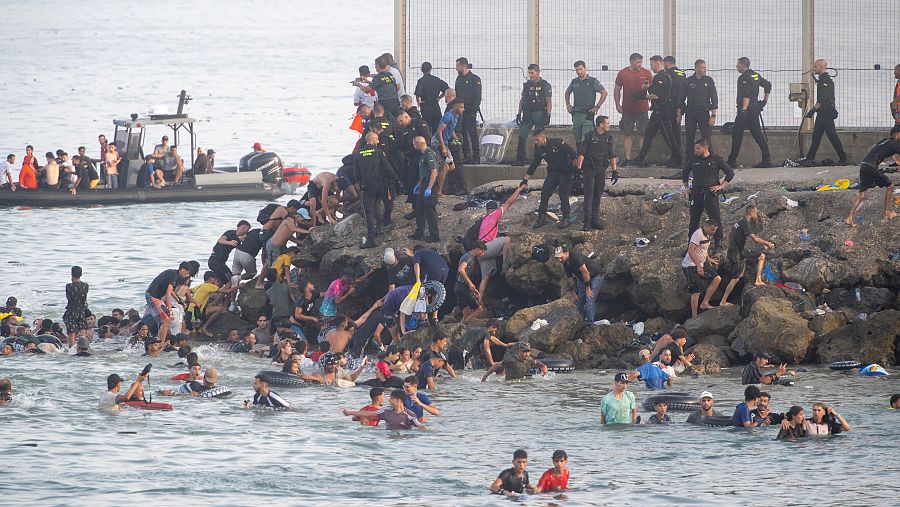 Multitud llega a costa, nadando y trepando rocas, asistida por Guardia Civil. Lancha semirrígida en el agua. Escena de urgencia y aglomeración.
