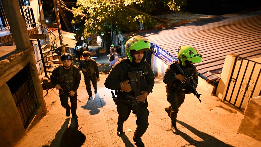 Policías con uniformes tácticos y armas largas patrullan una calle nocturna. Dos de ellos llevan cascos verdes brillantes, avanzando entre edificios y una valla ornamental.