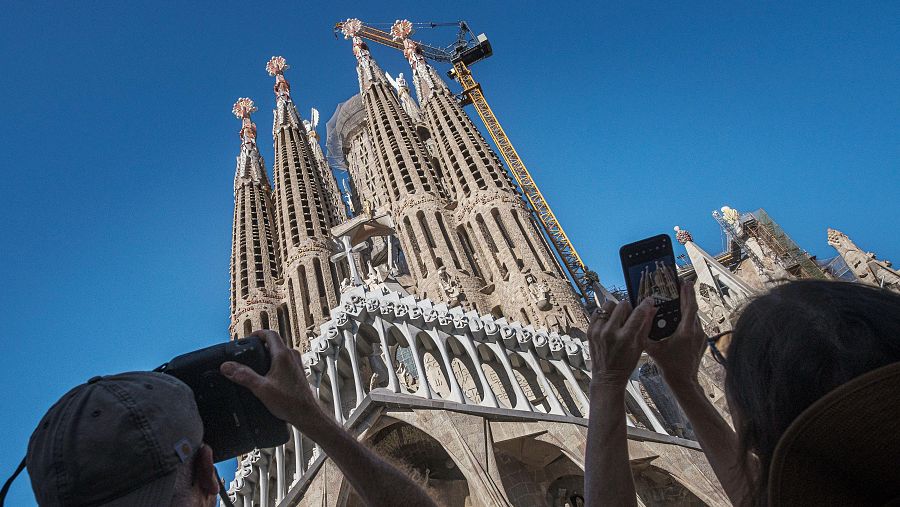 Vista de contrapicado de la Sagrada Familia con grúas de construcción. Dos personas, una con cámara y otra con móvil, la fotografían bajo un cielo azul.