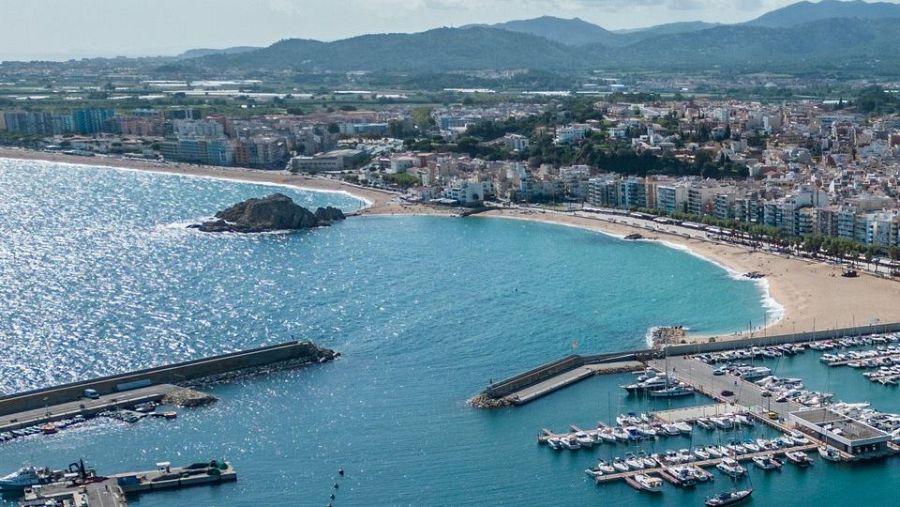 Vista aèria de la façana marítima de Blanes: badia turquesa, platges daurades, formació rocosa, port esportiu amb iots i ciutat densament edificada.