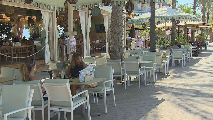 Terraza exterior con toldo a rayas verdes y blancas, mesas y sillas vacías. Dos mujeres sentadas, una lee un periódico. Se ve un cartel de 
