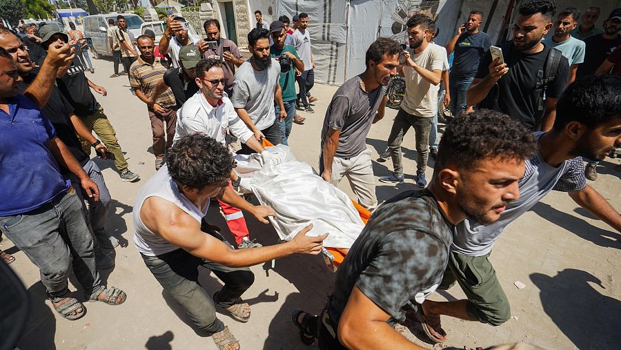 Hombres transportan un cuerpo en camilla de emergencia en Gaza, Palestina, el 19 de agosto de 2026. La escena es observada por una multitud que documenta el evento.