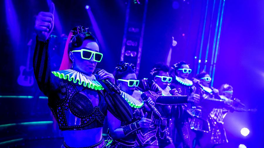 El cast del West End en una actuación, donde se puede ver la singularidad de la golilla fluorescente y la reinvención del vestuario barroco