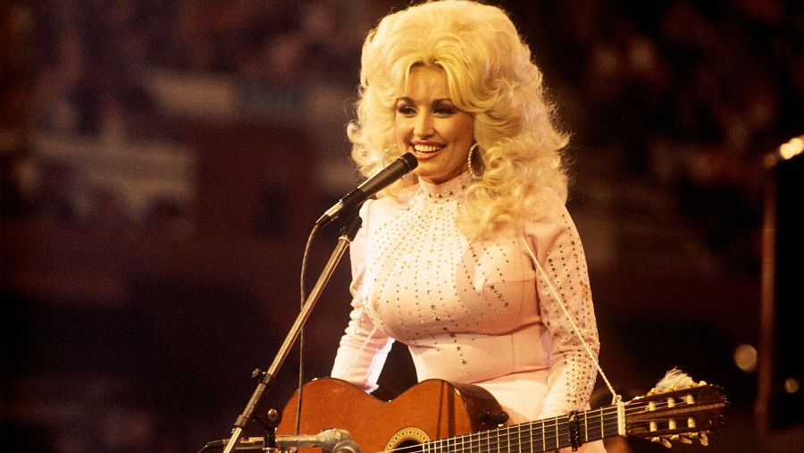 Dolly Parton, con su voluminoso cabello rubio y un brillante traje rosa, canta y toca la guitarra en 1976, iluminada en un escenario.
