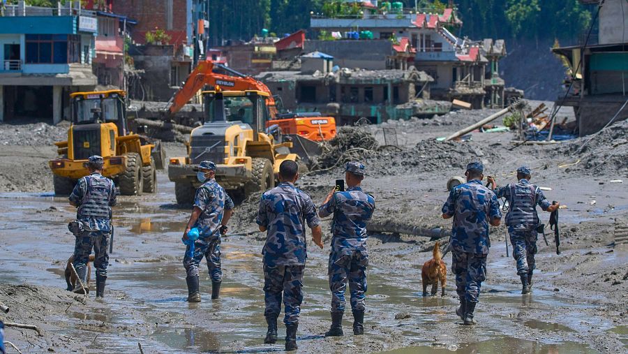 Personal de rescate en uniforme de camuflaje avanza por terreno fangoso y lleno de escombros, con maquinaria pesada trabajando y un pueblo devastado al fondo bajo un cielo despejado.