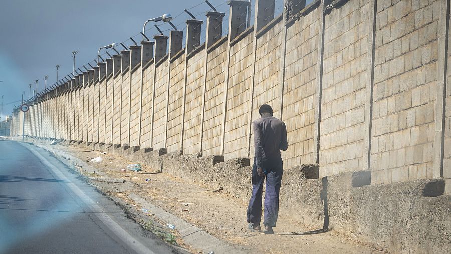 Persona de espaldas camina por camino de tierra junto a alto muro de hormigón con alambre de espino y farolas. Carretera asfaltada a la izquierda. Cielo nublado.