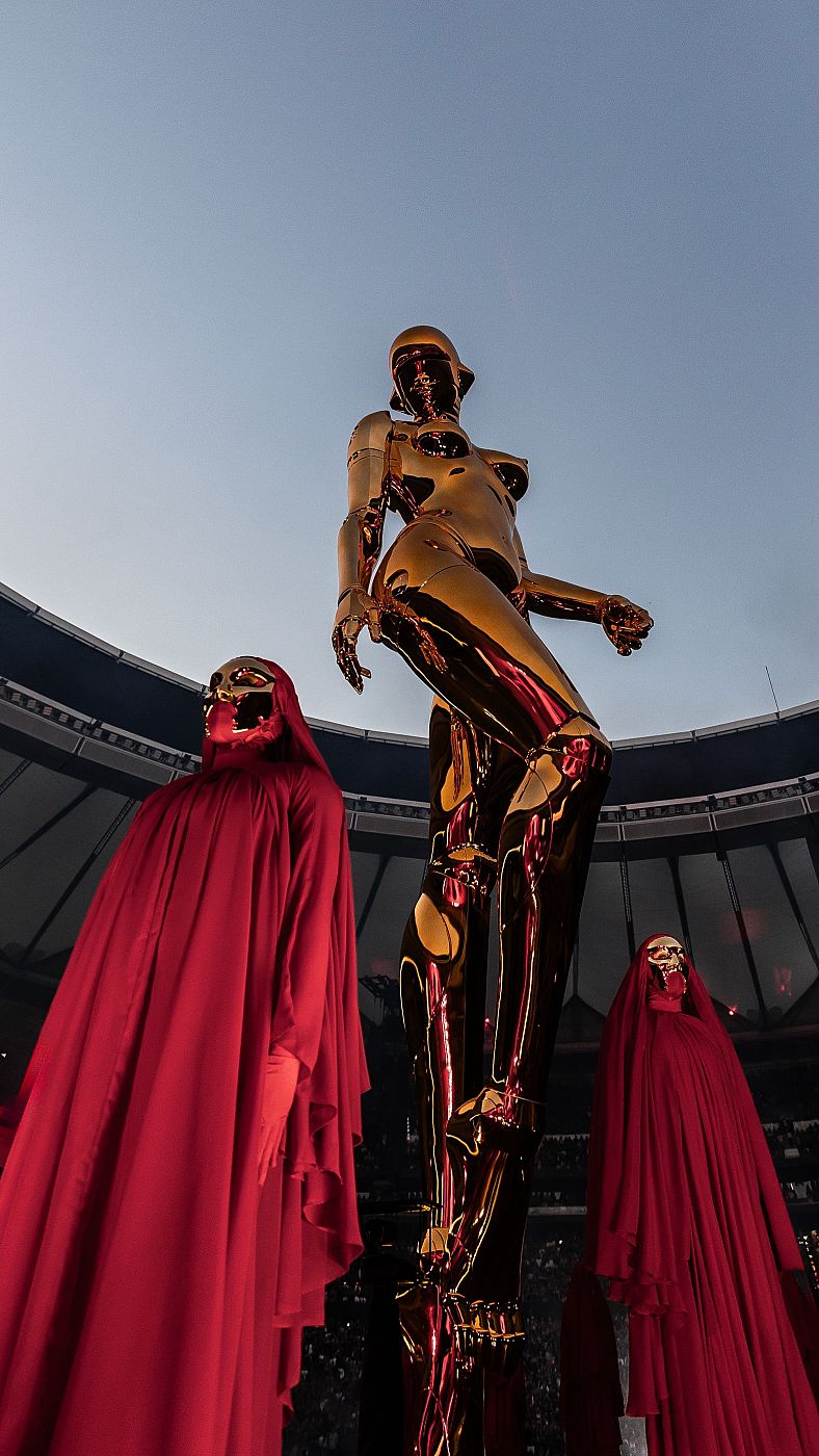 Escenario de concierto con estatua dorada futurista y figuras con túnicas rojas y máscaras doradas, en un estadio.
