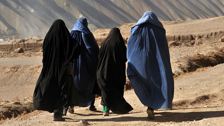 Cuatro mujeres con túnicas oscuras caminan por un camino de tierra en un paisaje árido y montañoso, con sombras alargadas.