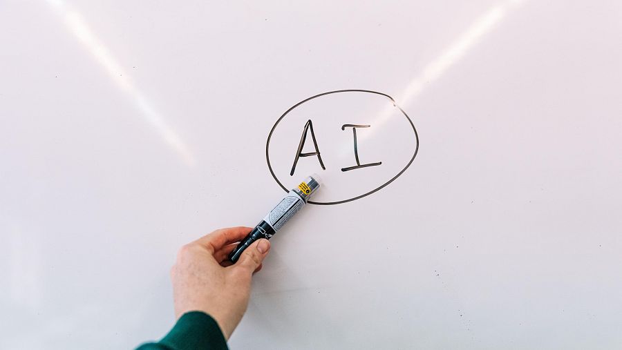 Hay una pizarra. Una mano señala con un bolígrafo las letras A I