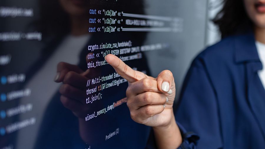 Mujer dando clase junto a un monitor colgado de una pared donde se ven lineas de código