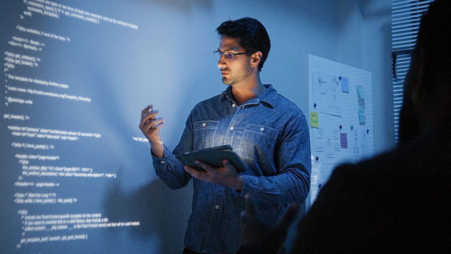 Profesor dando explicaciones. En la pared, una imagen proyectada de código de programación