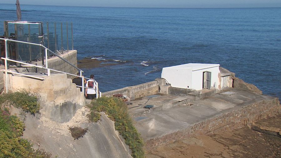 Persona desciende escaleras de hormigón con barandilla blanca hacia plataforma costera con edificio blanco y mar azul.