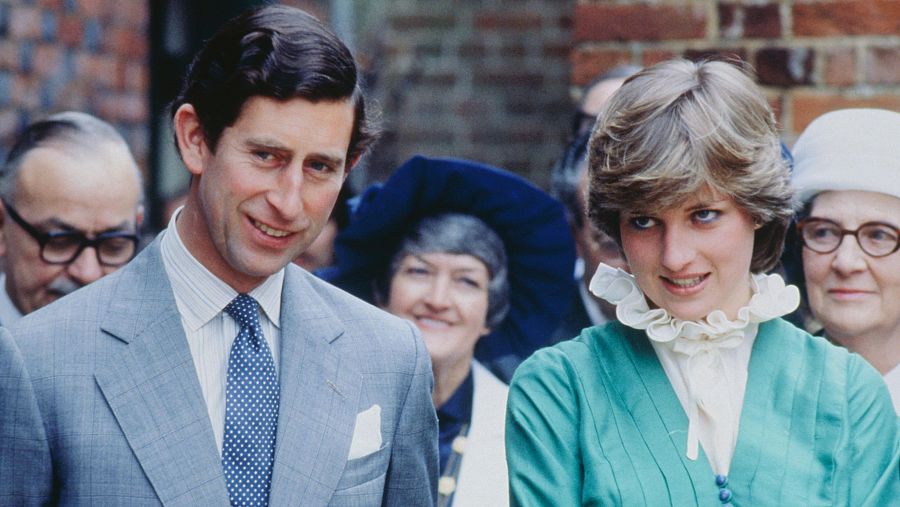 Primer plano del Príncipe Carlos y Lady Diana Spencer en un evento. Carlos con traje gris y corbata azul; Diana con vestido verde azulado y cuello blanco. Fondo de ladrillo y público difuminado.