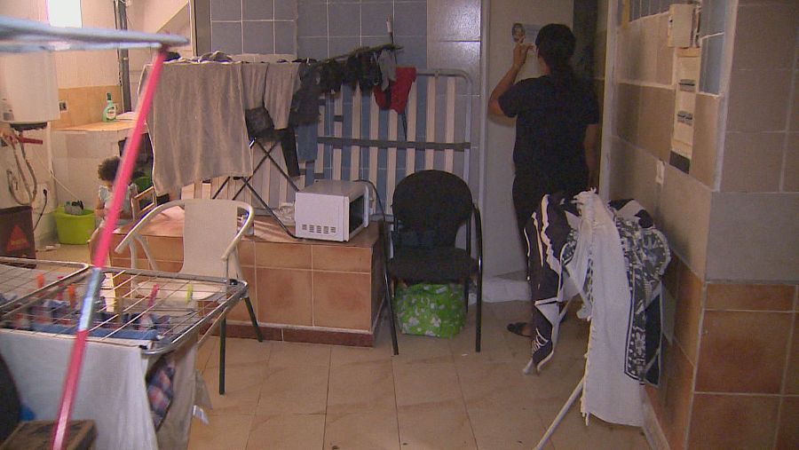 Interior doméstico con tendederos, un adulto de espaldas, un niño/a cerca de un cubo verde y mobiliario funcional. Paredes de azulejos y suelo de baldosas.