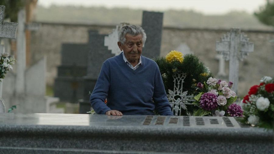 Hipólito, de cabello canoso y tez clara, se muestra pensativo en un cementerio, con flores a su lado y lápidas al fondo.