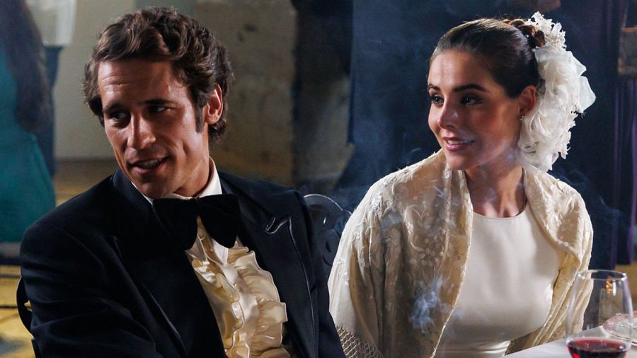 Martiño Rivas y Carmen Fernández Raigón, vestidos de gala, posan sonrientes en un evento formal. Él con esmoquin, ella con vestido blanco y tocado floral.