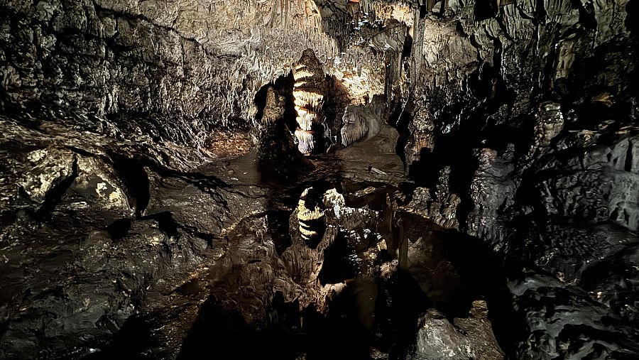 Interior de cueva con estalactitas, estalagmitas y una columna central iluminada. Tonos oscuros y contrastes dramáticos. Sin personas ni texto visible.