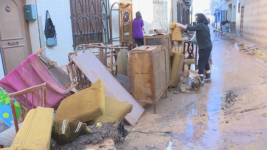 Calle residencial anegada por lodo con muebles arruinados y una mujer limpiando tras las inundaciones.