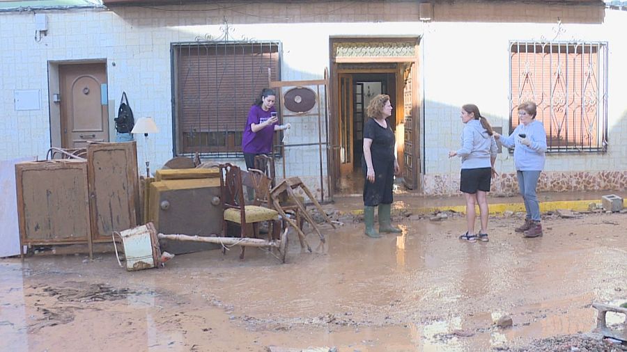 Calle residencial inundada con muebles dañados en el lodo y cuatro mujeres conversando frente a una casa afectada.