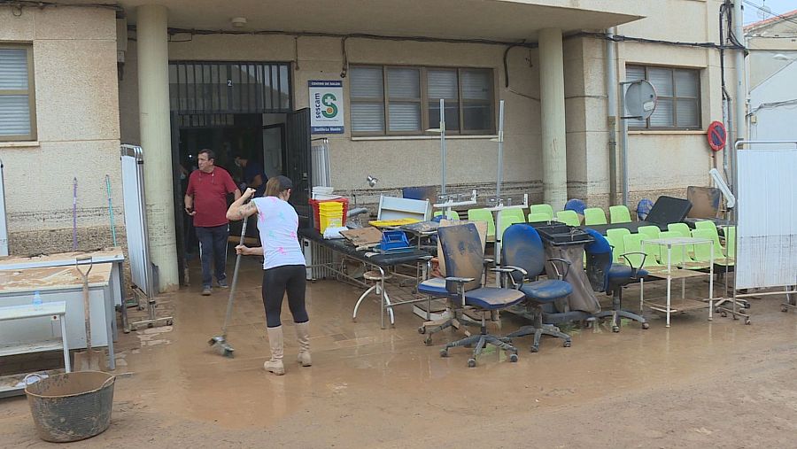 Exterior de un centro de salud inundado de lodo donde una mujer con botas de agua limpia el suelo y se acumulan muebles y material clínico manchados.