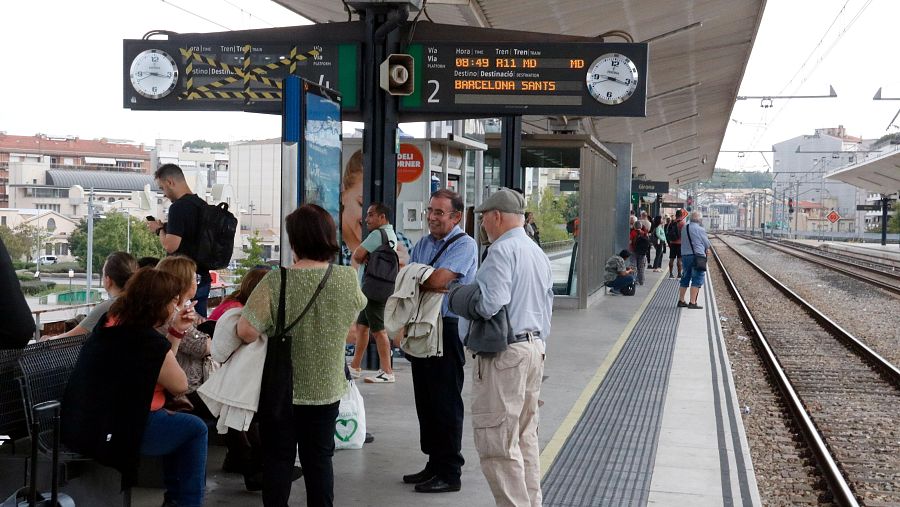 Passatgers esperant a l'andana d'una estació de tren sota una coberta metàl·lica, amb panells informatius i edificis al fons.