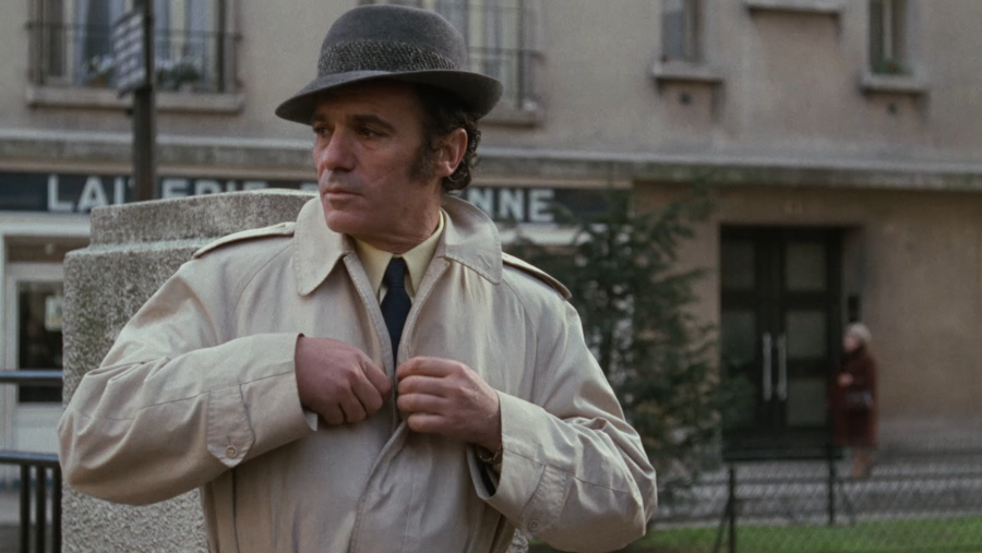 Plano medio de Paco Rabal de perfil con gabardina beige, sombrero gris y mirada seria en una calle clásica.