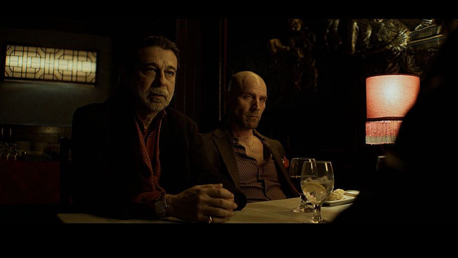 Dos hombres con semblante serio, vestidos con chaqueta, están sentados a una mesa de restaurante con iluminación tenue y una lámpara encendida.
