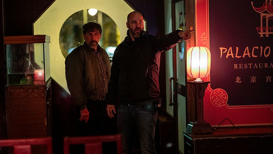 El director Albert Pintó y el actor Luis Zahera en un set de rodaje ambientado en un restaurante oriental con iluminación tenue y detalles de atrezo.