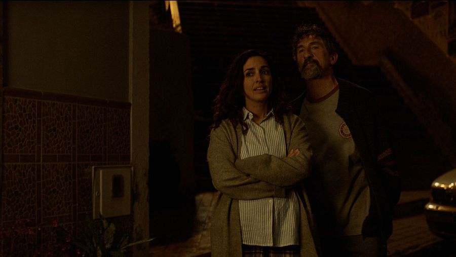Plano medio nocturno de los actores Inma Cuesta y Luis Zahera de pie en el exterior, mirando serios hacia fuera de campo con iluminación tenue.