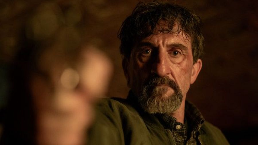 Primer plano del actor Luis Zahera con expresión severa, barba perilla y chaqueta verde militar, apuntando con un arma.