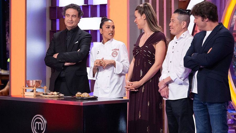 Cinco personas posan sonrientes tras una mesa de cata con platos de degustación en el plató de un programa de cocina con iluminación anaranjada y violeta.