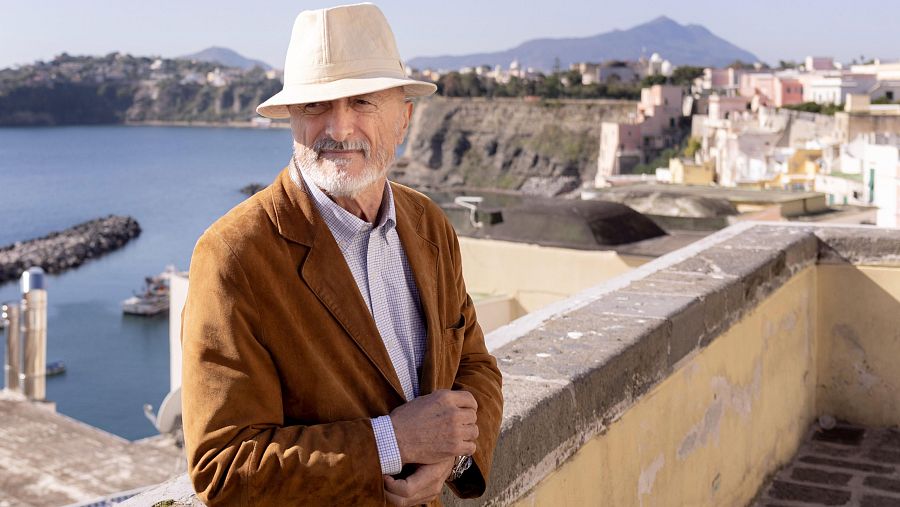 Plano medio del escritor Arturo Pérez-Reverte con sombrero beige y chaqueta marrón en una terraza con vistas al mar y paisaje costero.